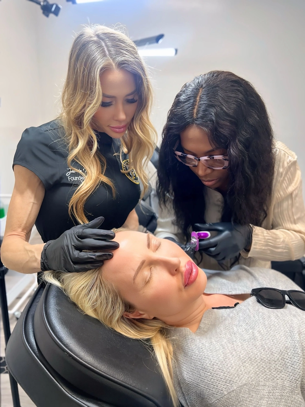 Two women wearing gloves perform a scalp treatment on a blonde woman reclining in a chair.