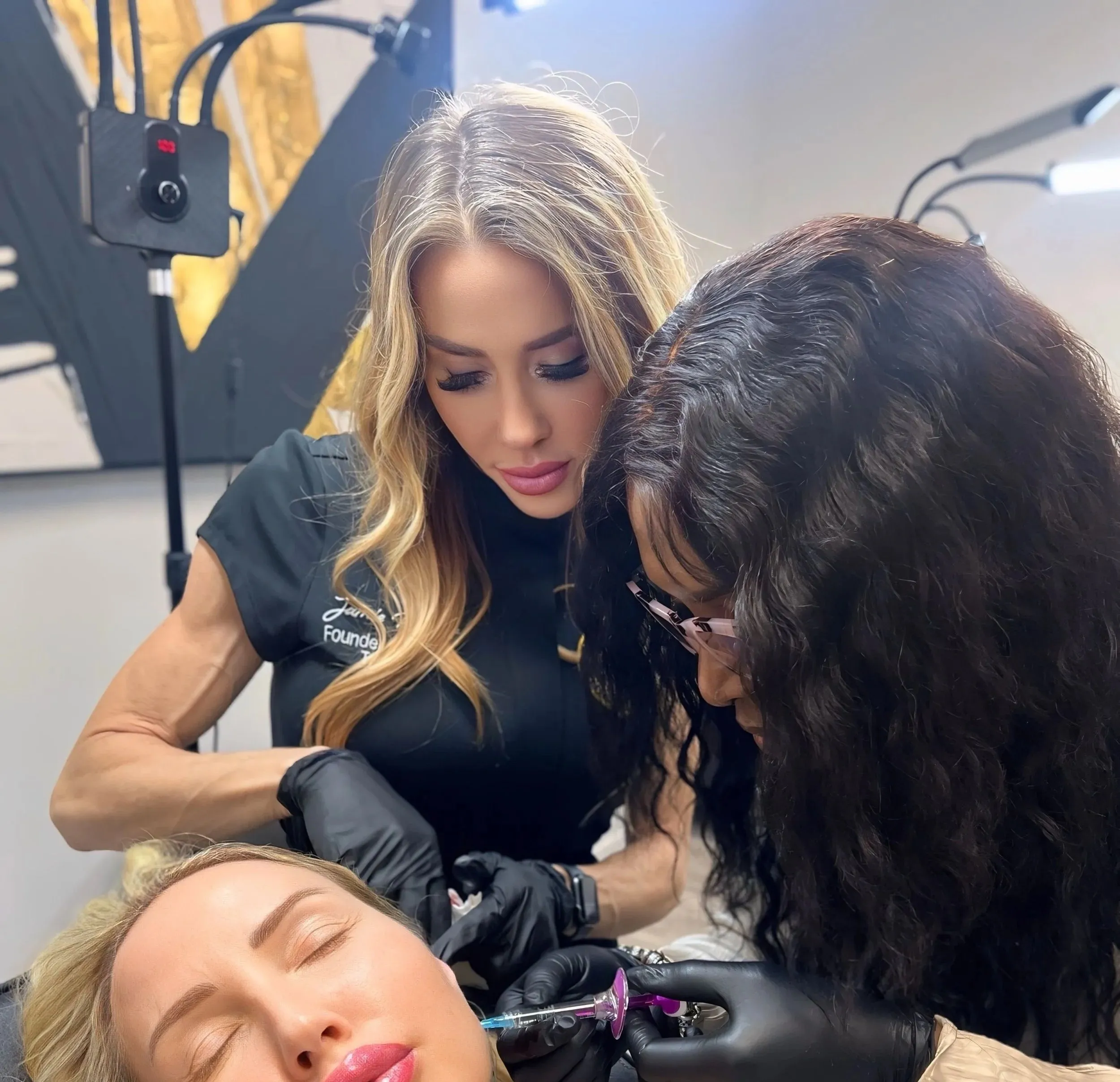 A woman receives a cosmetic injection in her face from two professionals wearing black gloves.