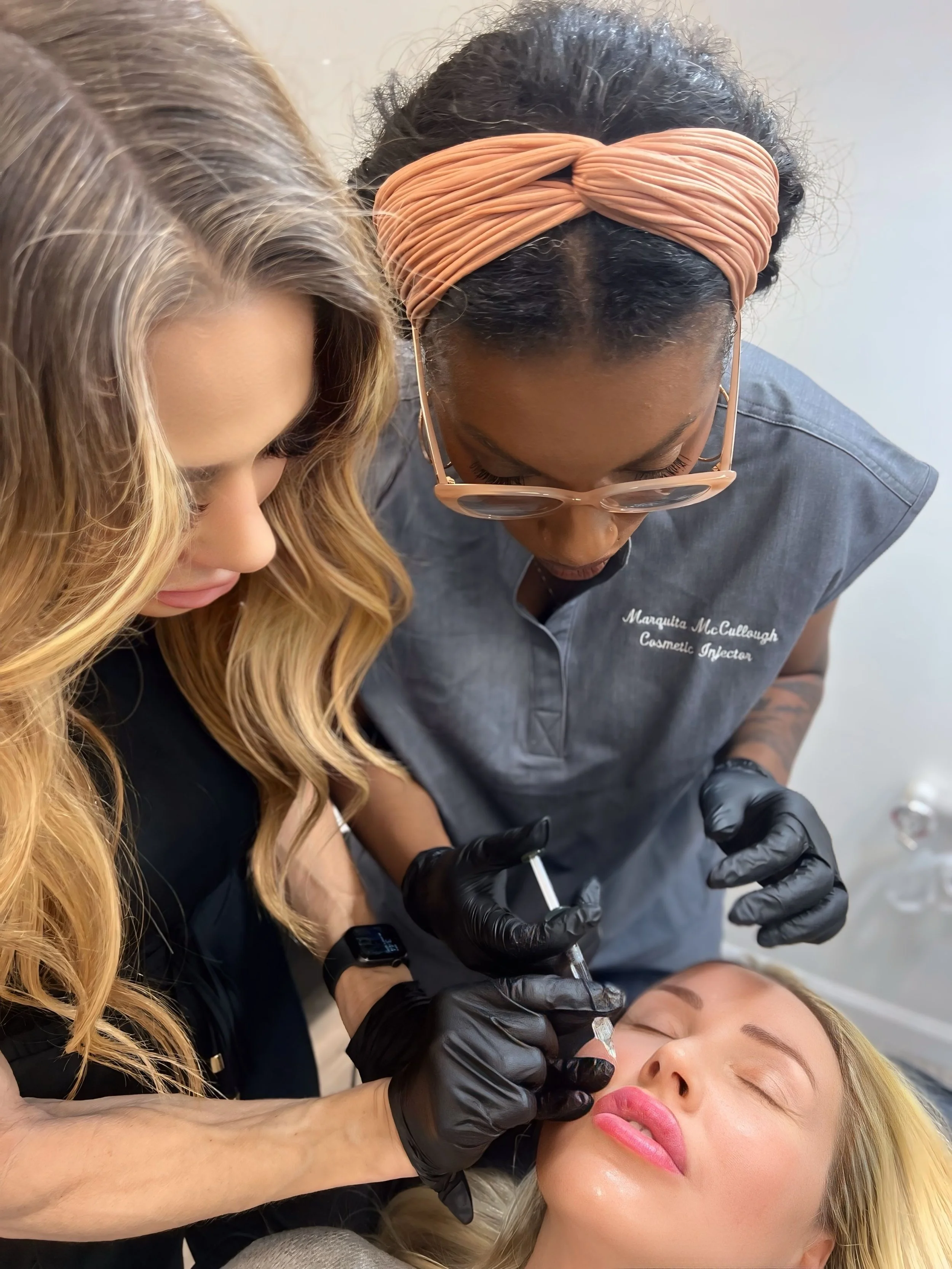 Cosmetic injector wearing gloves injects a woman’s lips while another watches closely.