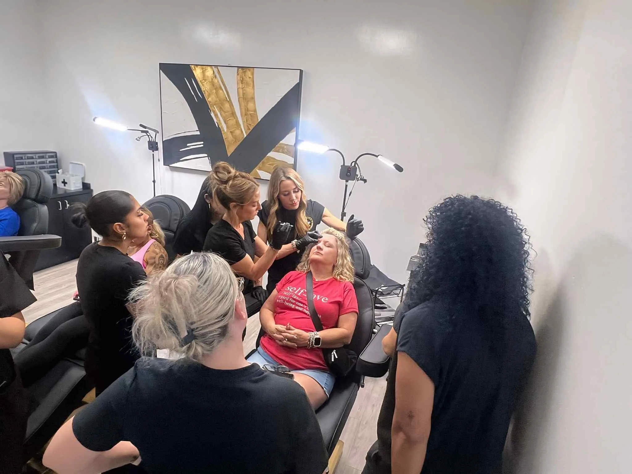 A woman in a salon chair receives a cosmetic eyebrow procedure while others observe closely.