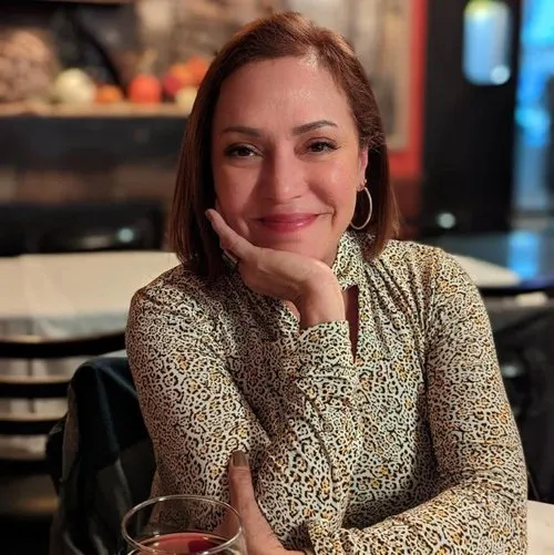 Smiling woman with shoulder-length hair in a patterned top sitting at a table with a drink.
