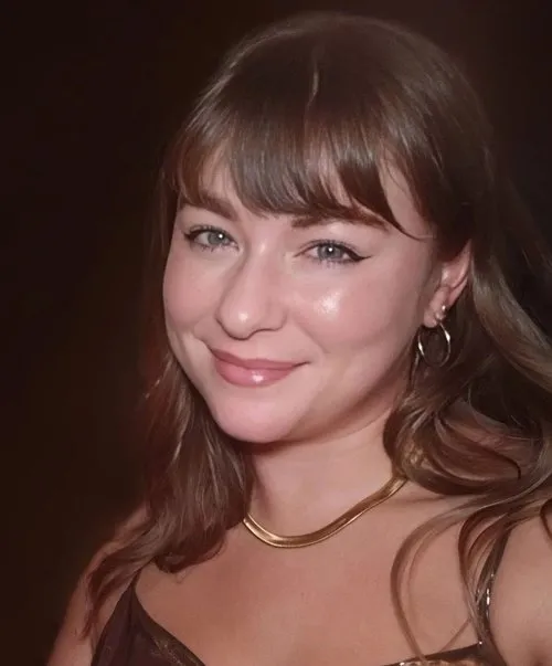 Smiling young woman with brown hair, hoop earrings, and a gold necklace against a dark background.