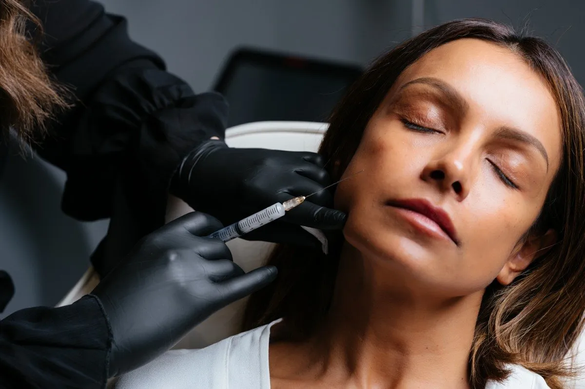 Woman with eyes closed receiving a facial injection from a person wearing black gloves.