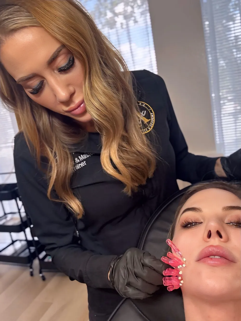 A woman with blonde hair wearing black gloves holds a pink facial massage tool on another woman's cheek.