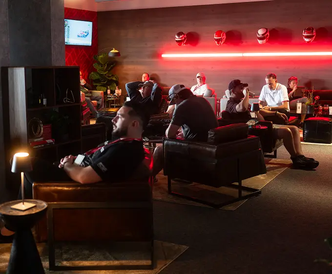 Men sitting on black leather chairs in a dimly lit room with red lighting and racing helmets on the wall.