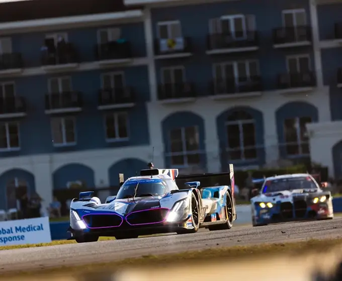 Two race cars on a track with building and spectators in the background under the sun.
