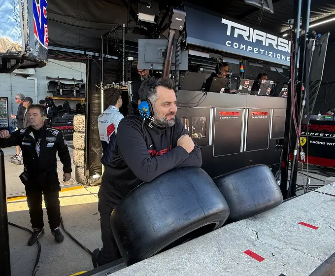 Man with headset leaning on two large racing tires at a racing team pit area with others working nearby.