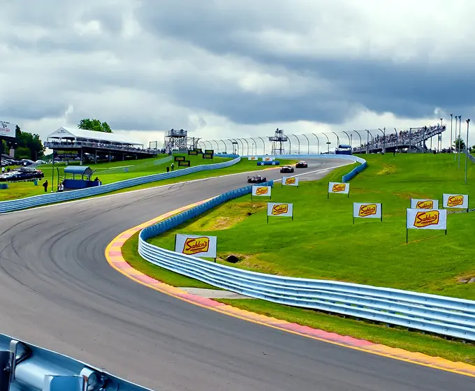 Race cars speeding on a curved track with green grass and spectator stands under a cloudy sky.