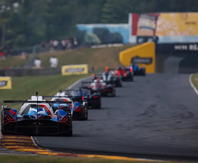 Race cars lined up on a track with a blurred audience and barriers in the background.