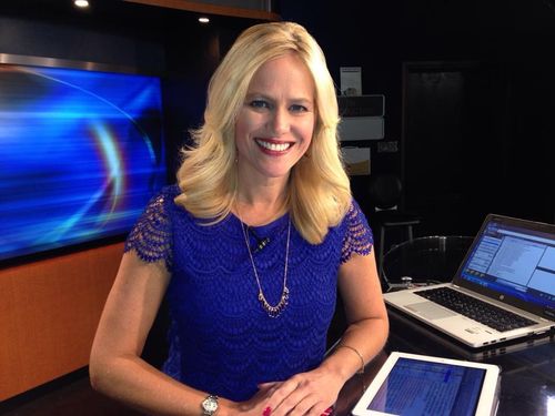 Lynda Halligan in a blue lace top sitting at a news desk with a laptop and tablet, smiling at camera.