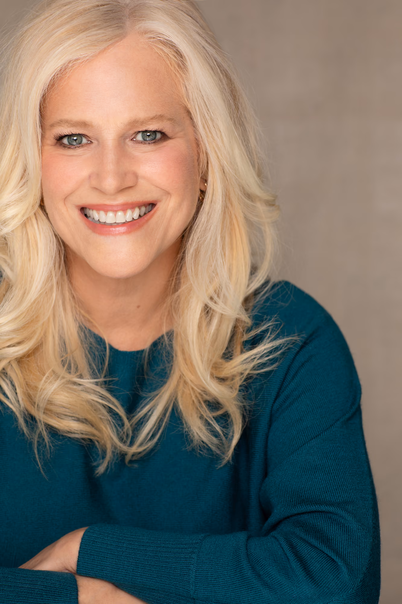 Smiling blonde woman (Lynda Halligan)with blue-green eyes wearing a teal sweater against a beige background.