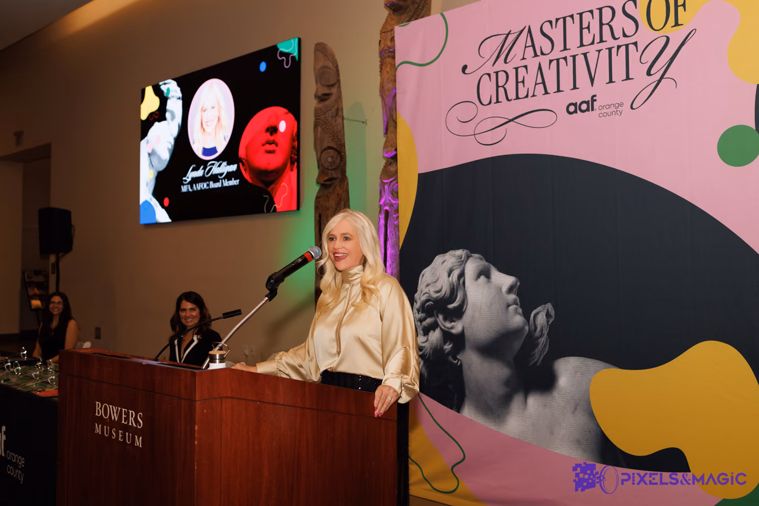 Lynda Halligan speaking at podium at Bowers Museum with Masters of Creativity backdrop and seated attendees in background.