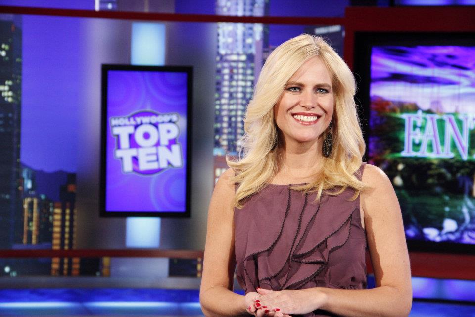 Lynda Halligan in purple dress stands in TV studio with 'Hollywood's Top Ten' sign in background.