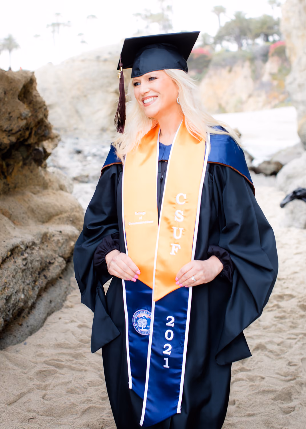 Lynda Halligan wearing black cap and gown with CSUF 2021 stole on a sandy beach.