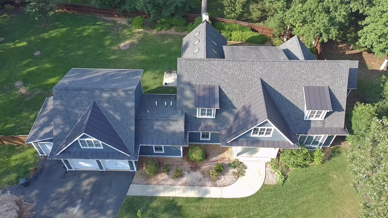 Aerial view of a large gray home with a complex roofline, black metal awnings, attached garage, driveway, and fenced backyard.
