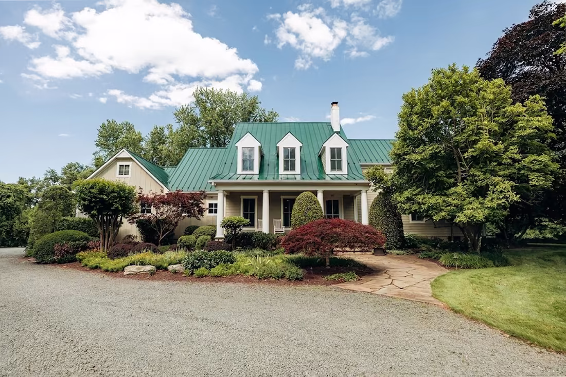 Traditional cream-colored home with a green metal roof, dormer windows, covered front porch, and mature landscaping.