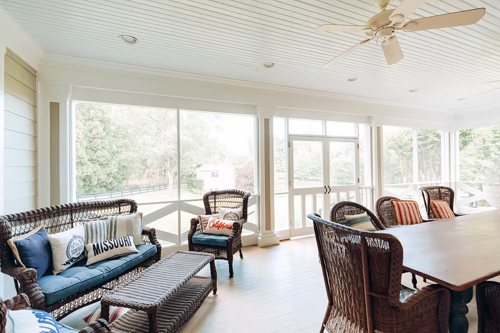 Bright enclosed porch with white beadboard ceiling, wicker furniture, dining table, and large screened windows.