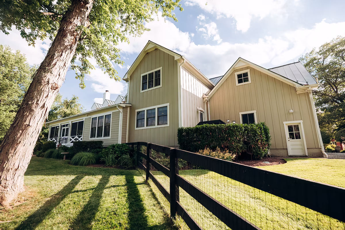Large beige board-and-batten home with multiple gables, black metal roof accents, screened porch, and black fence.