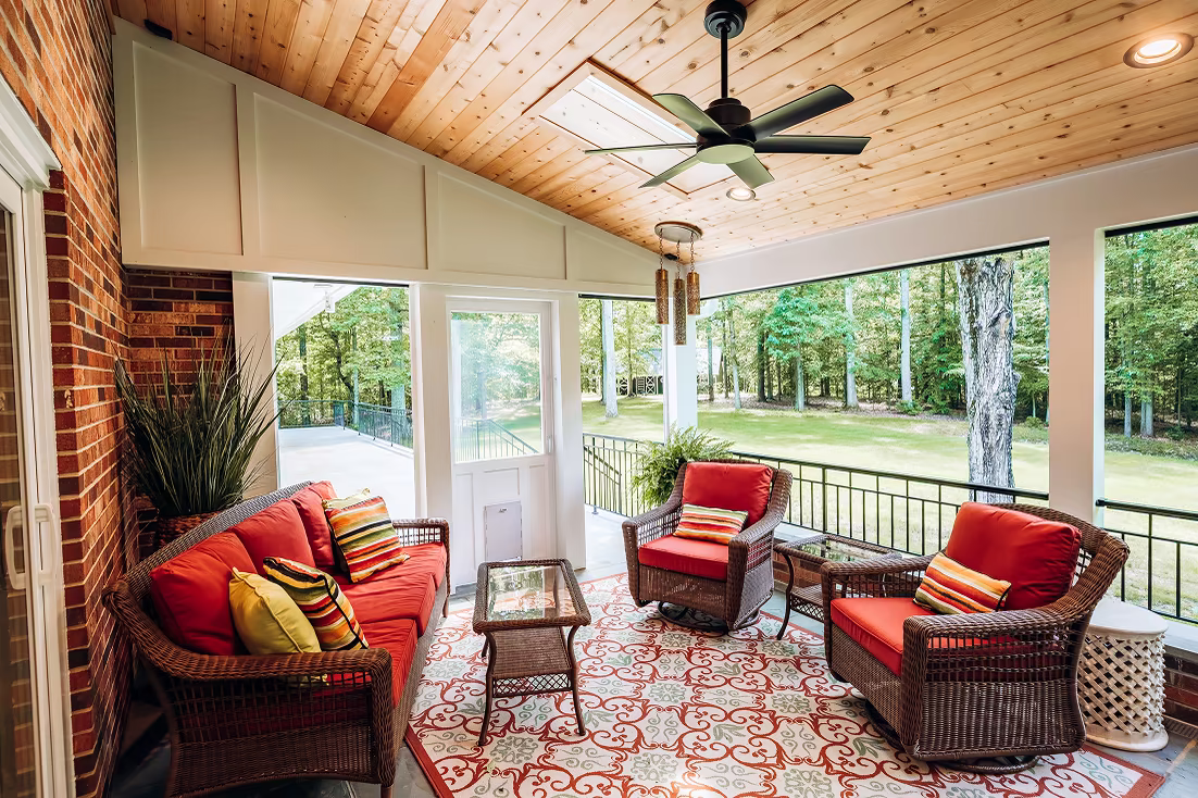 Screened porch with a wood-paneled ceiling, ceiling fan, red wicker seating, and views of a wooded backyard.