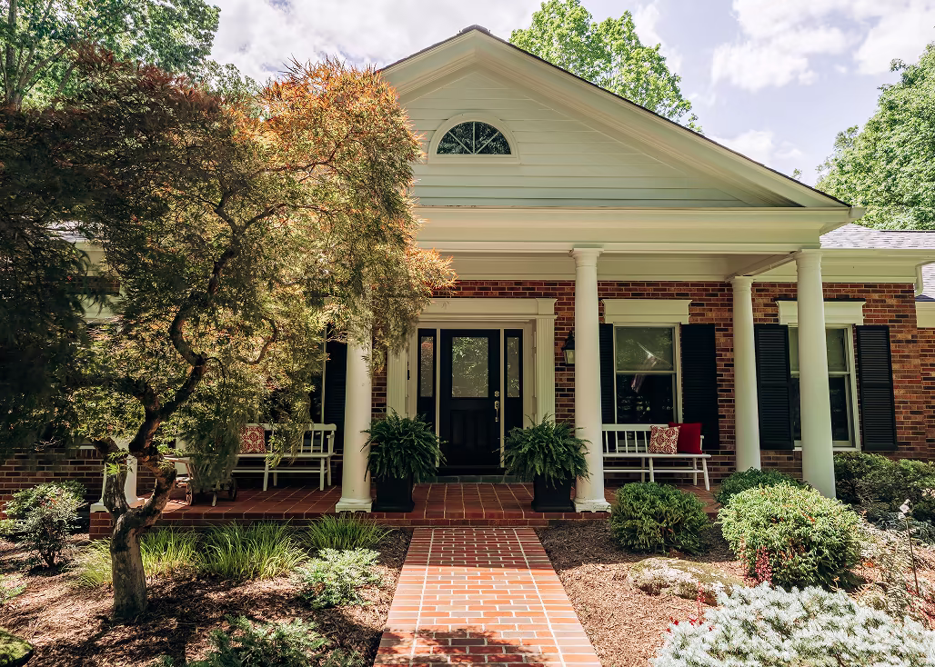 Brick colonial-style home with white columns, a covered front porch, black shutters, and landscaped front walk.
