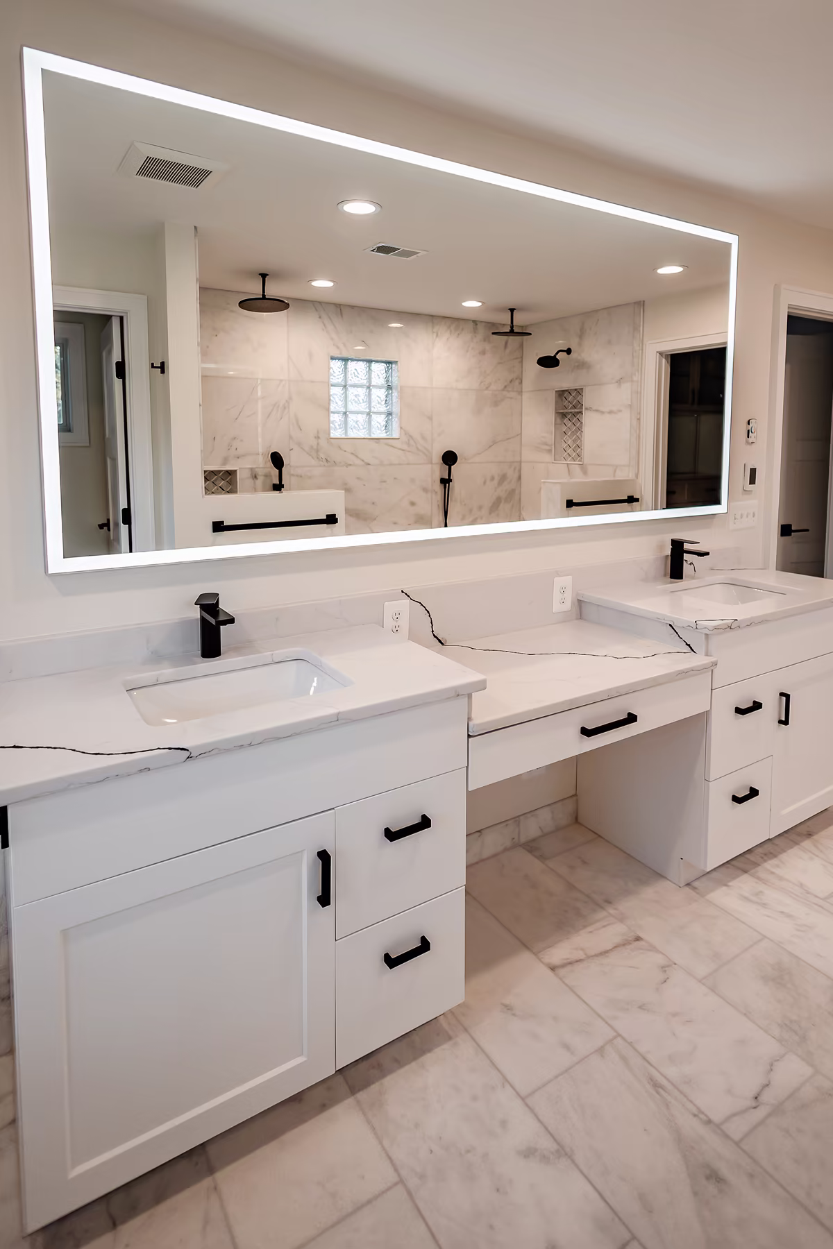 Double white vanity with marble countertops, black hardware and faucets, illuminated wall mirror, and marble walk-in shower in the reflection.