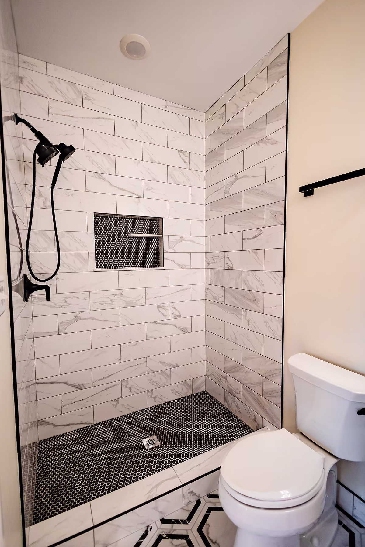 Compact walk-in shower with marble-look subway tile walls, black penny tile floor, black shower fixtures, and adjacent white toilet.