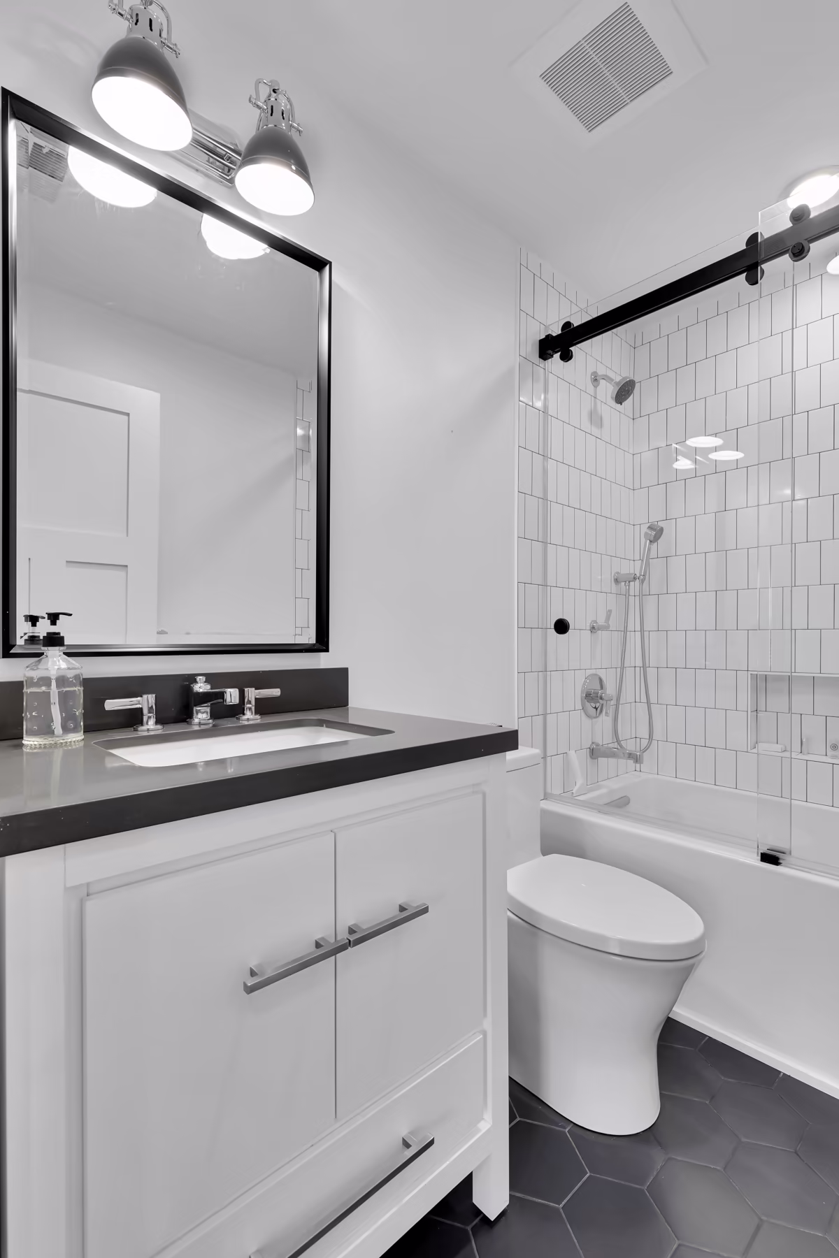 Modern white bathroom with a black countertop vanity, black-framed mirror, white subway tile tub surround, and dark hexagon floor tile.