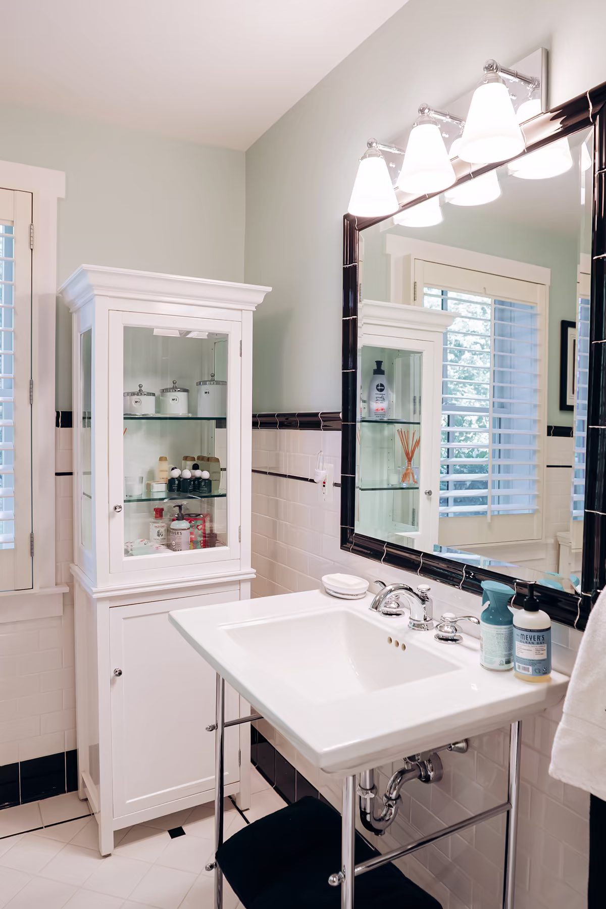 Classic bathroom vanity with a white pedestal-style sink, black-framed mirror, chrome fixtures, glass display cabinet, and white subway tile walls.