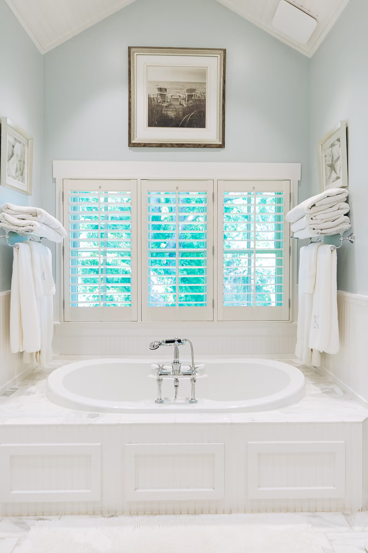 Bright traditional bathroom with an oval soaking tub, white plantation shutters, pale blue walls, marble surround, and folded white towels.