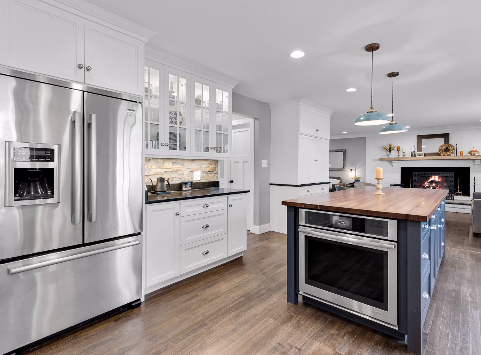 Open white-and-navy kitchen with stainless-steel appliances, a butcher-block island, pendant lights, and a fireplace visible in the adjoining living area.