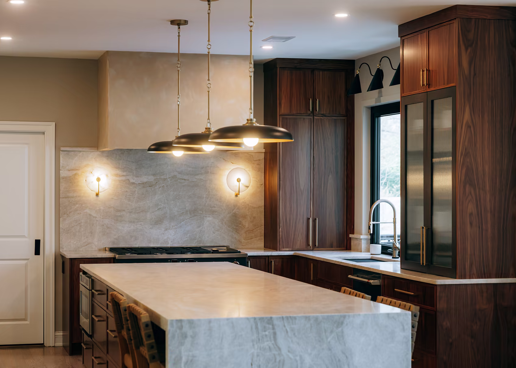 Contemporary kitchen with rich walnut cabinetry, a waterfall marble island, brass-and-black pendant lighting, and a marble backsplash.