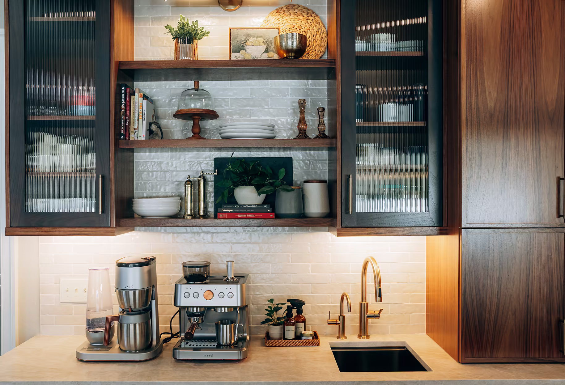 Dark walnut kitchen cabinetry with fluted-glass doors frames open shelving above a coffee station and brass sink fixtures.