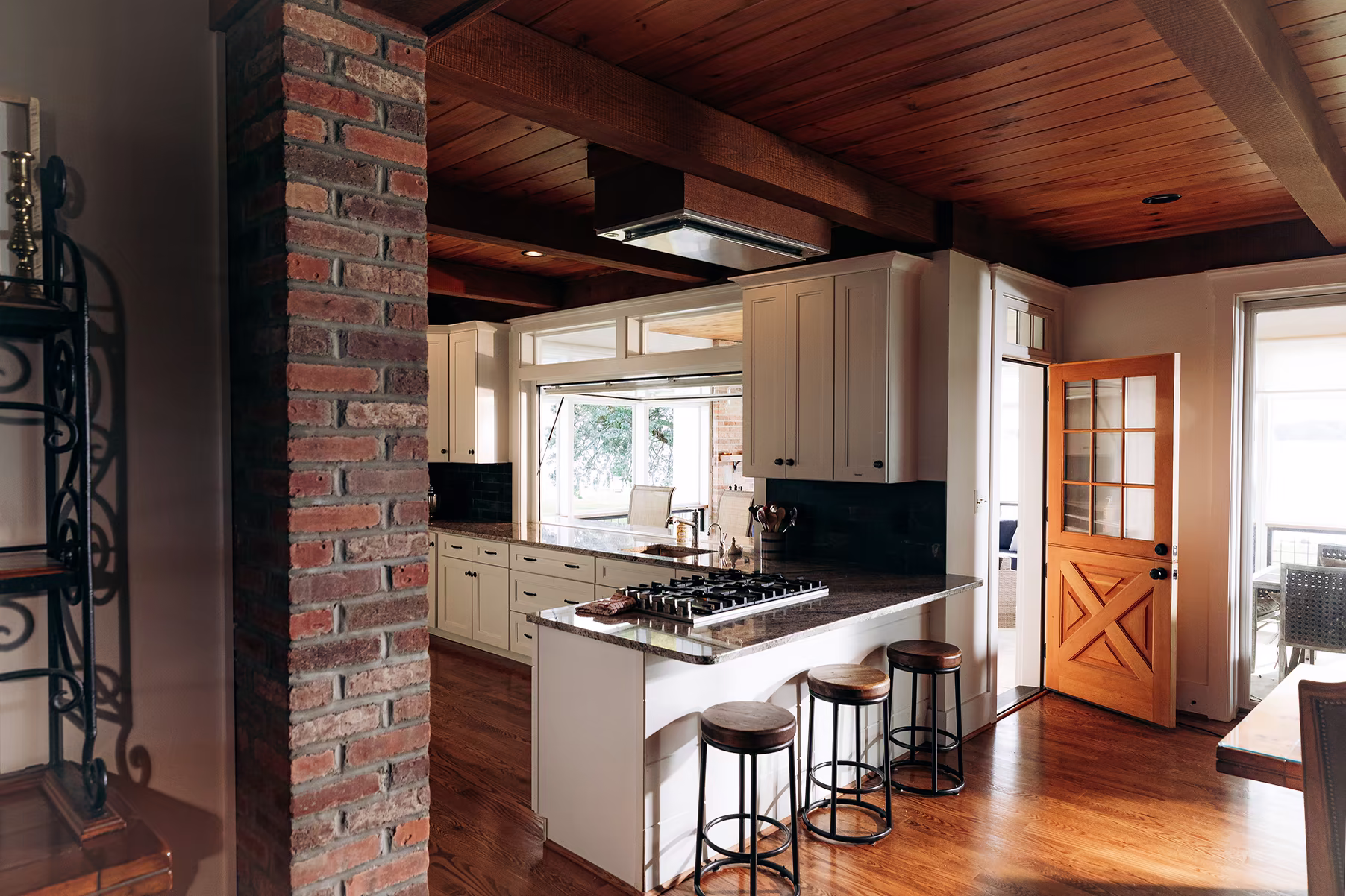 Traditional white kitchen with a granite-topped island, exposed brick column, dark wood ceiling beams, and four counter stools.