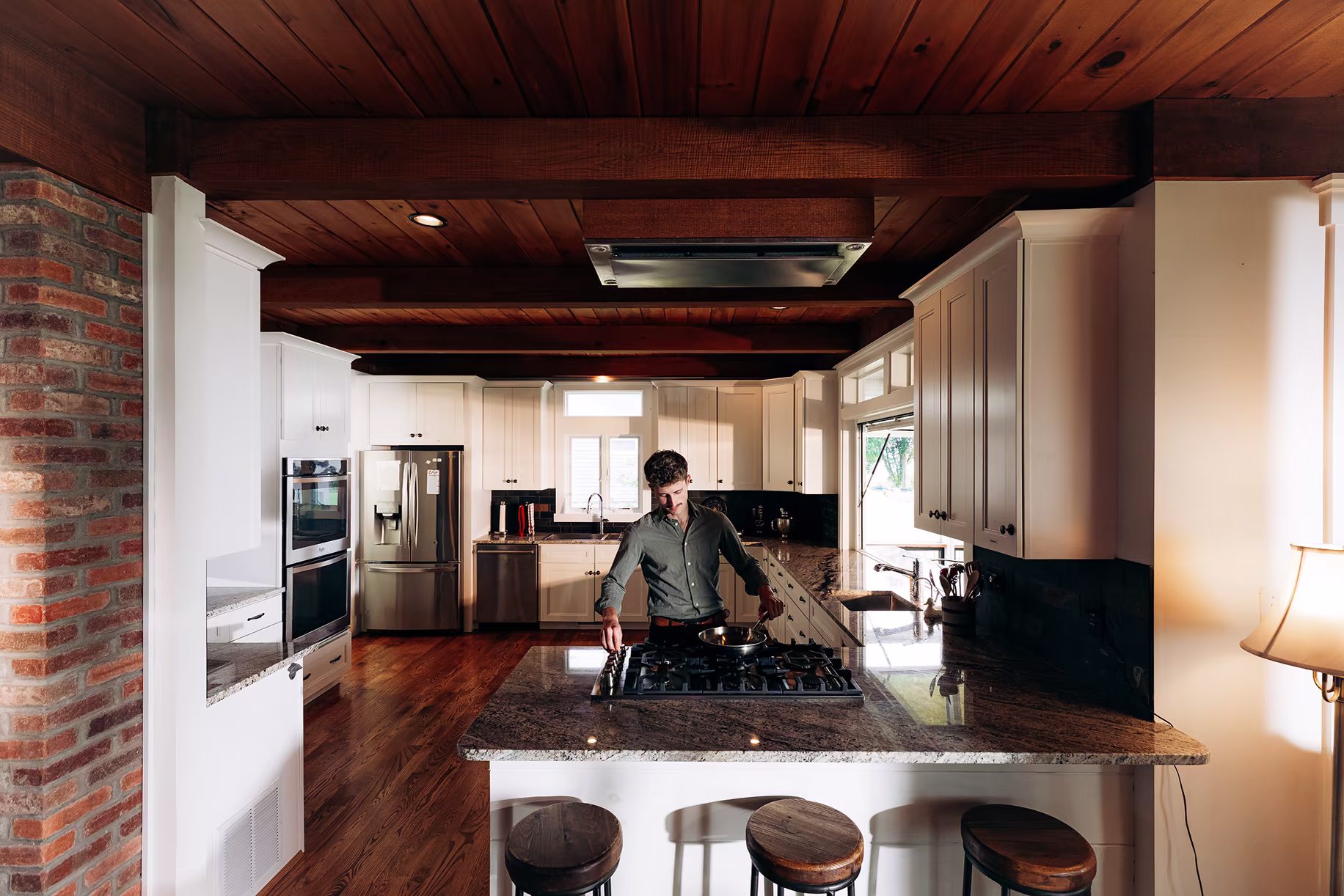 A man cooks at a large granite island with a gas cooktop in a traditional kitchen featuring white cabinetry, dark wood ceiling beams, exposed brick, and three wooden bar stools.