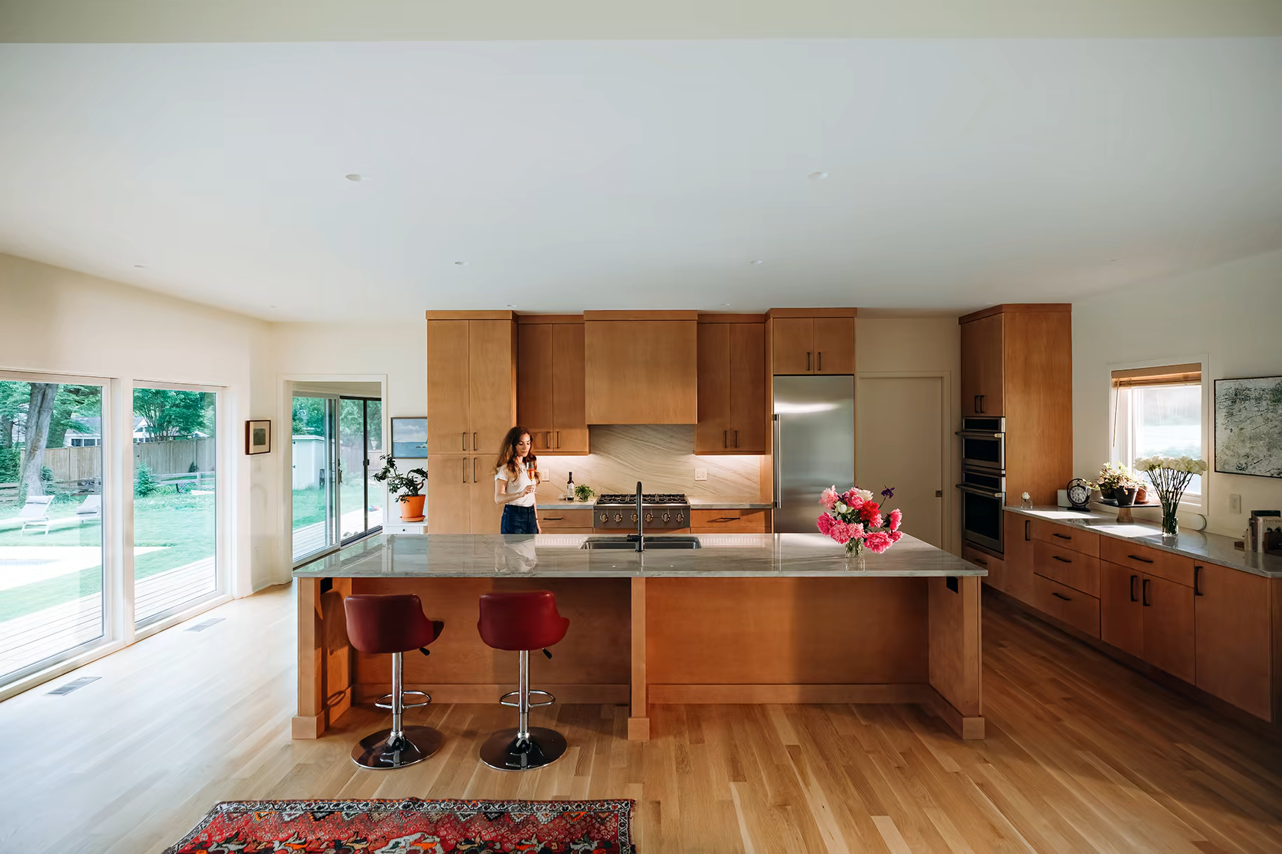 A bright, modern kitchen with warm wood cabinetry, a large marble-topped island, two red bar stools, and floor-to-ceiling windows looking onto a green backyard.