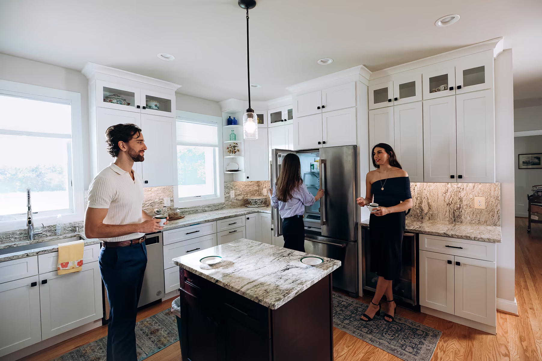Three people gather in a bright white kitchen with granite countertops, a central island, and stainless-steel appliances; one person opens the refrigerator while two others hold cups.