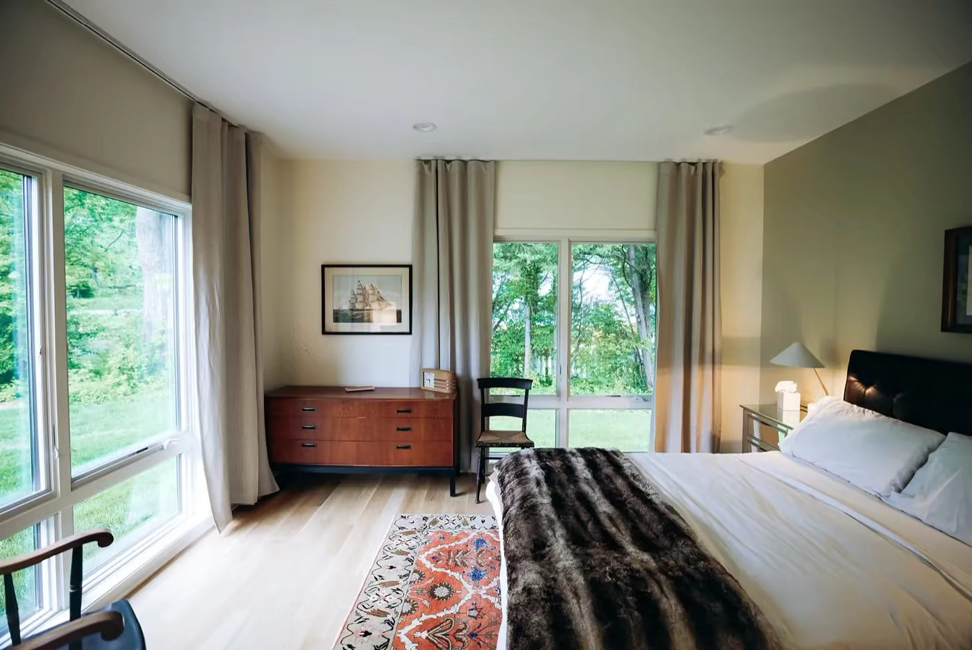 Primary bedroom with a fur throw at the foot of the bed, a wood dresser, and floor-to-ceiling windows overlooking the yard