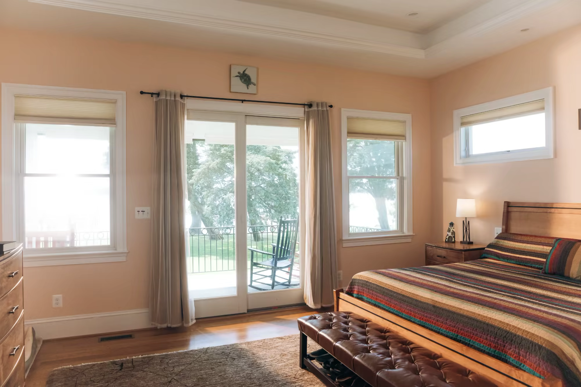 Peach-toned primary bedroom with a striped bedspread, leather bench, and French doors opening to a lakeside balcony