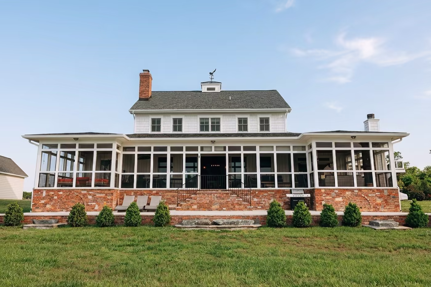 White cottage-style home with a wraparound screened porch, brick foundation, and a rooftop weathervane