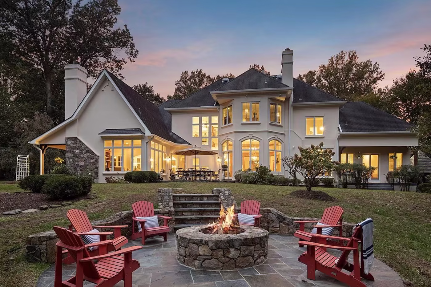Large stucco and stone home lit at dusk with a stone fire pit and red Adirondack chairs on the back patio