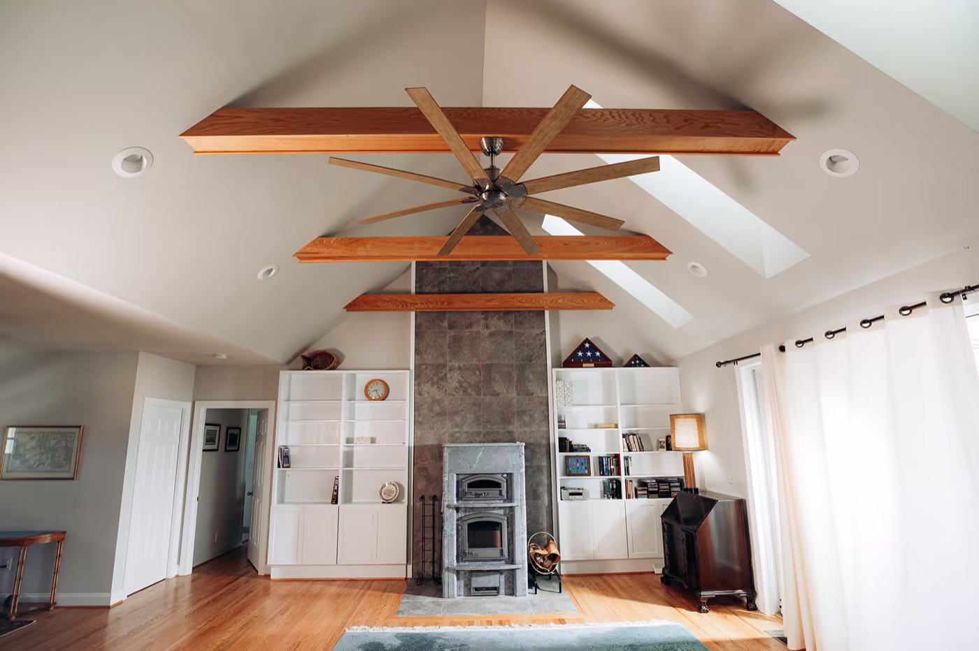 Living room with a vaulted wood-beamed ceiling, ceiling fan, stone fireplace surround, and white built-in shelving
