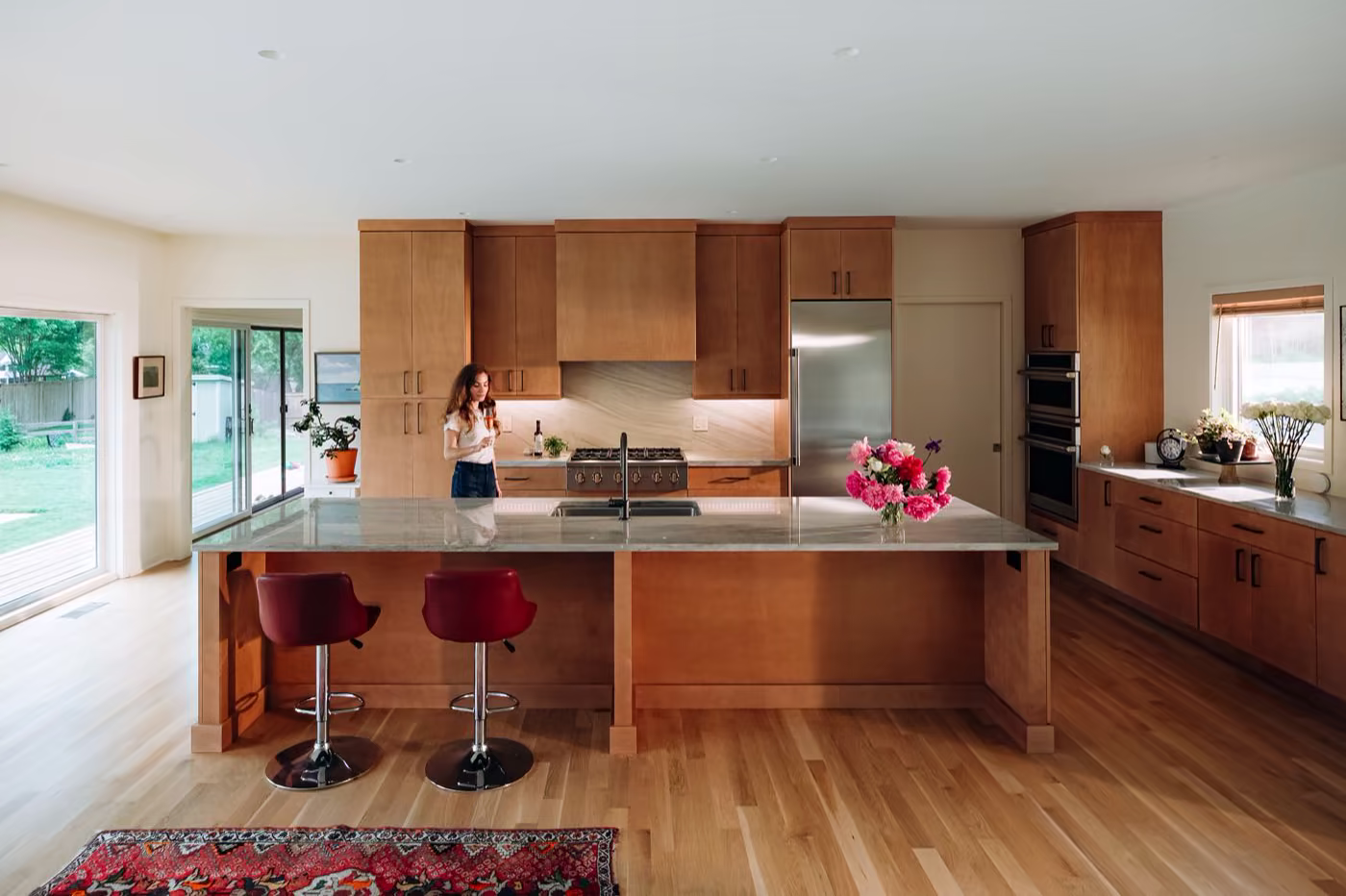 Kitchen with a large quartz island, wood cabinetry, stainless steel appliances, red counter stools, and a person cooking at the range