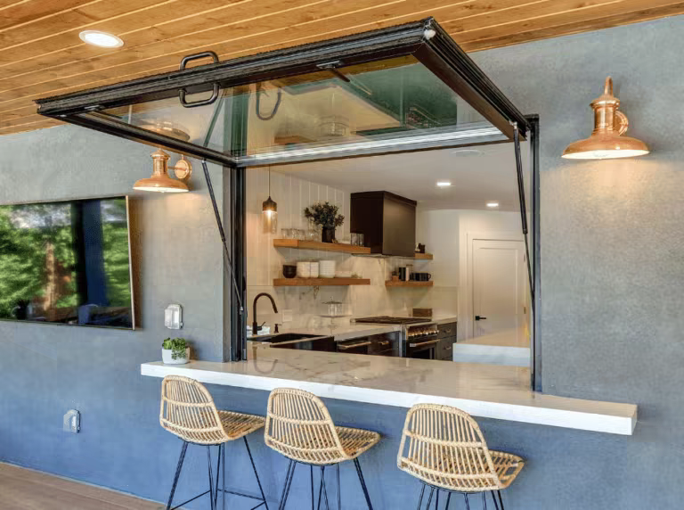 Outdoor bar with a large glass bi-fold window opened to a modern kitchen, showing pendant lighting and rattan barstools.