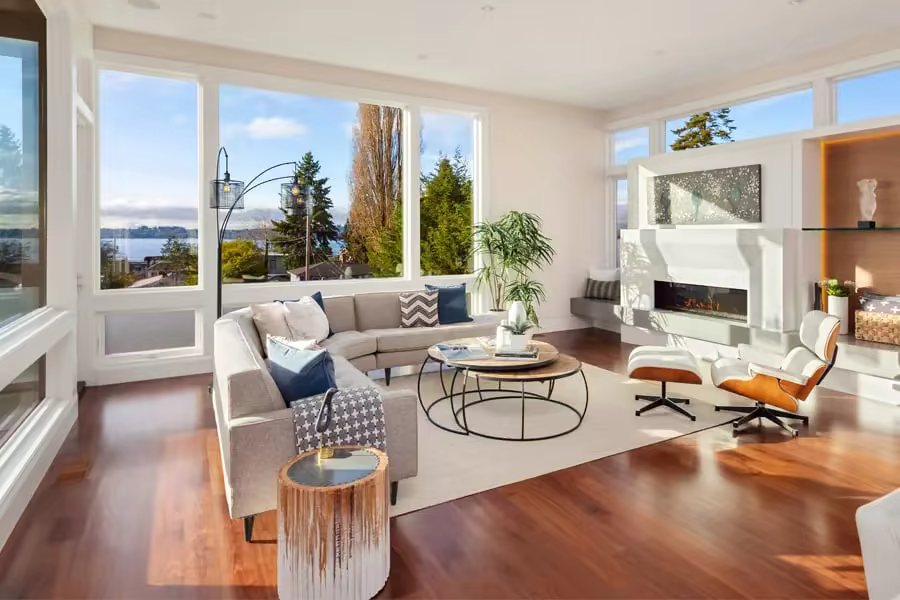 Living room with floor-to-ceiling windows overlooking a lake, wood flooring, and a fireplace seating area.