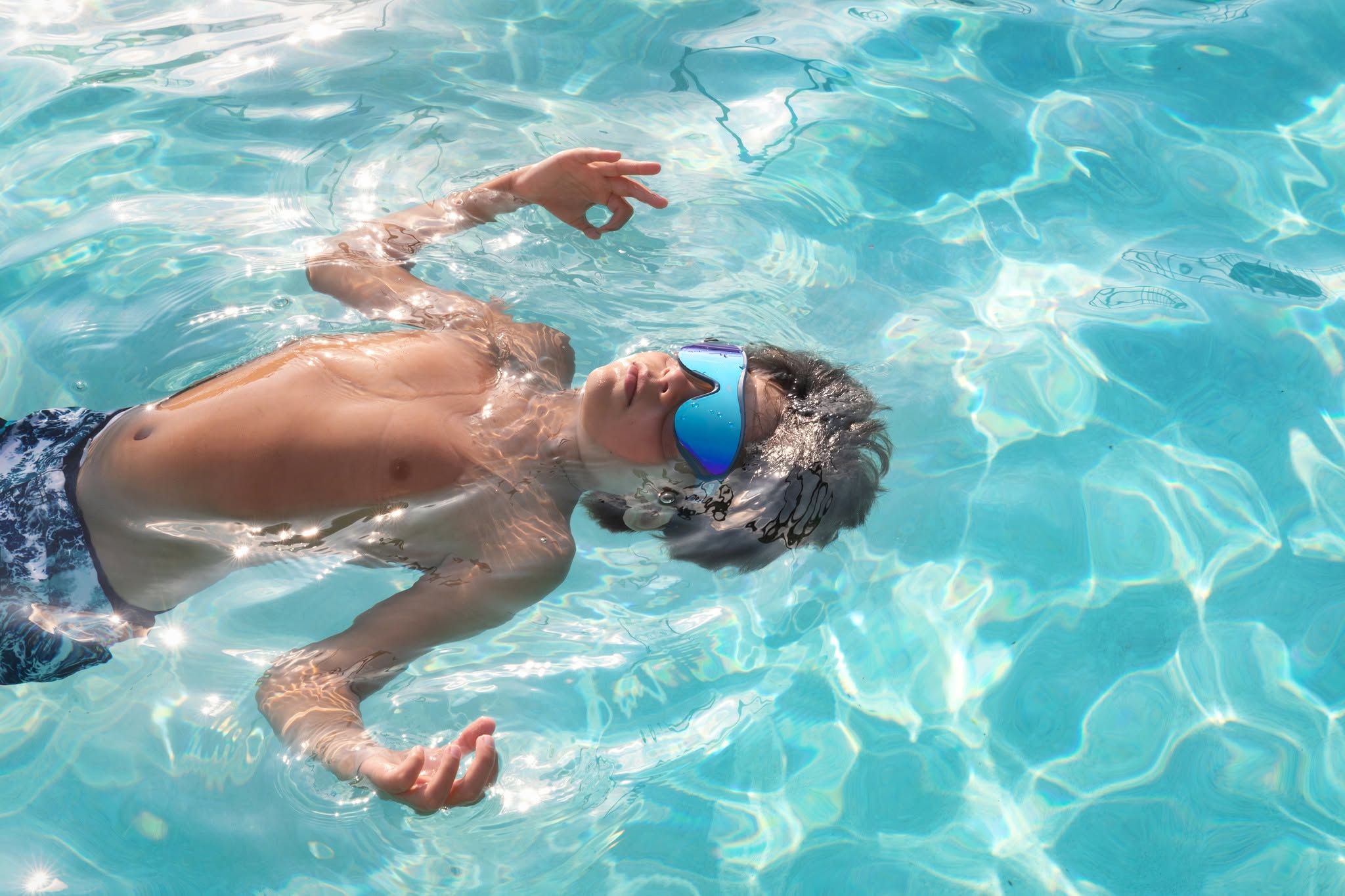 Boy wearing mirrored swim goggles floats peacefully in a sunlit swimming pool