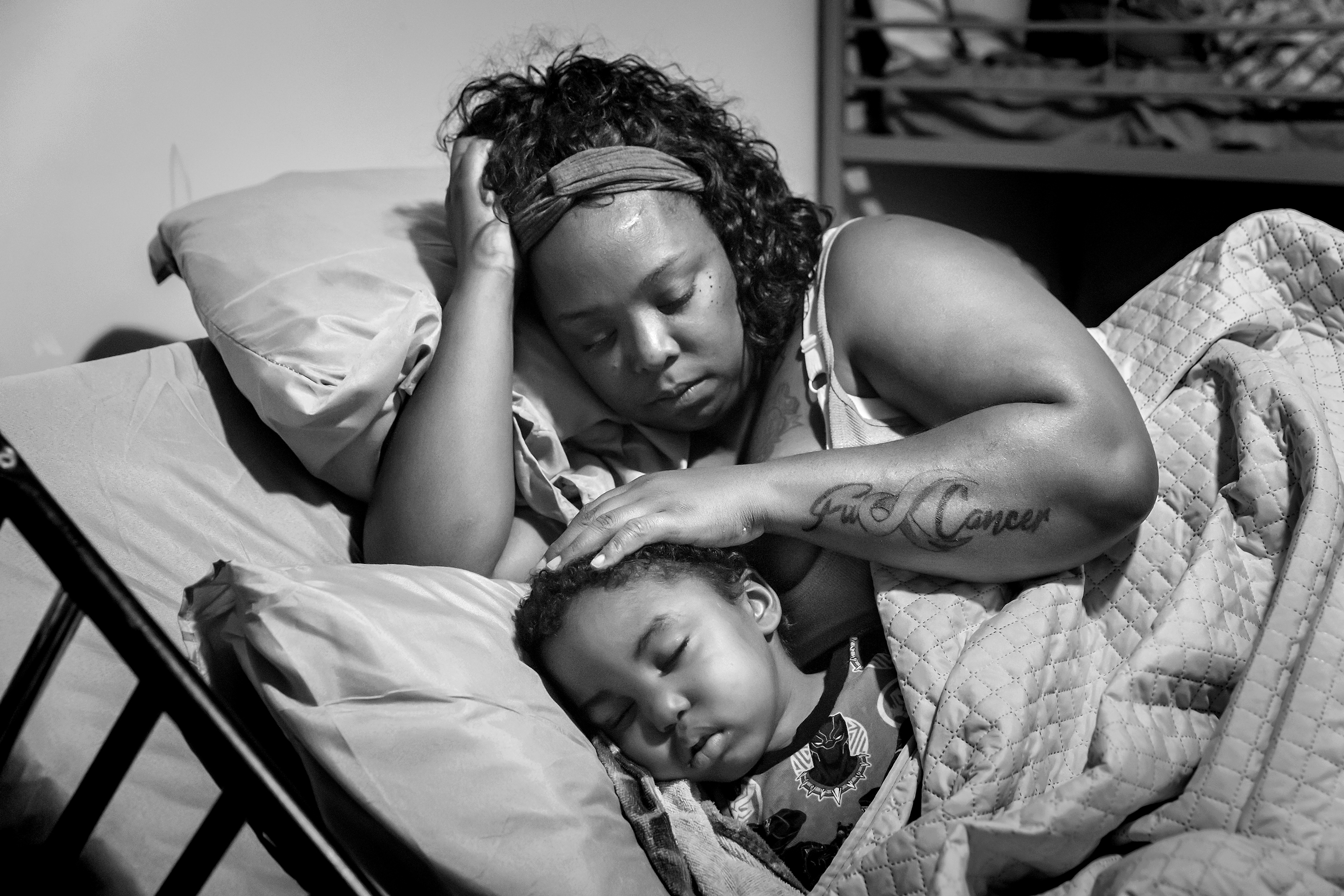 Black-and-white photograph of a mother resting beside her sleeping child in a hospital bed, with a visible forearm tattoo reading "F U Cancer."