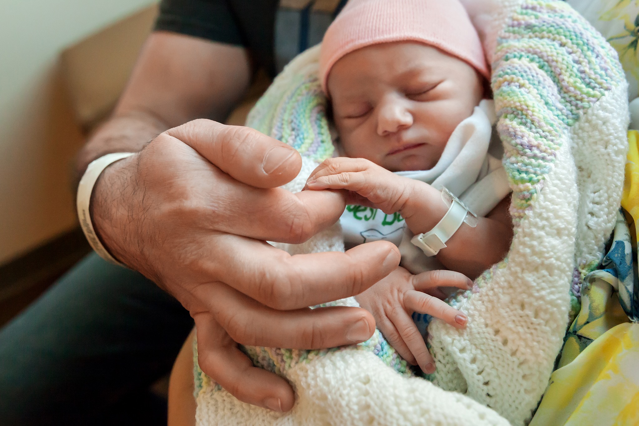 Close-up of a newborn wrapped in a knitted blanket, their tiny body nestled in their father's large hands while grasping his finger.