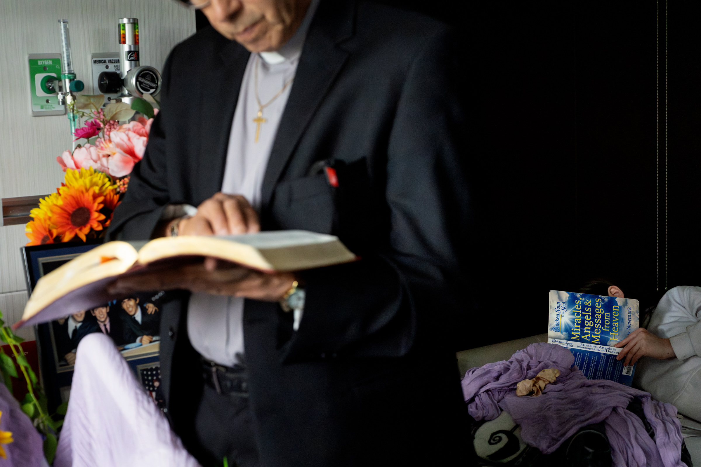 Hospital chaplain administers the Last Rites at a patient's bedside while a child quietly reads Chicken Soup for the Soul: Miracles, Angels & Messages from Heaven.