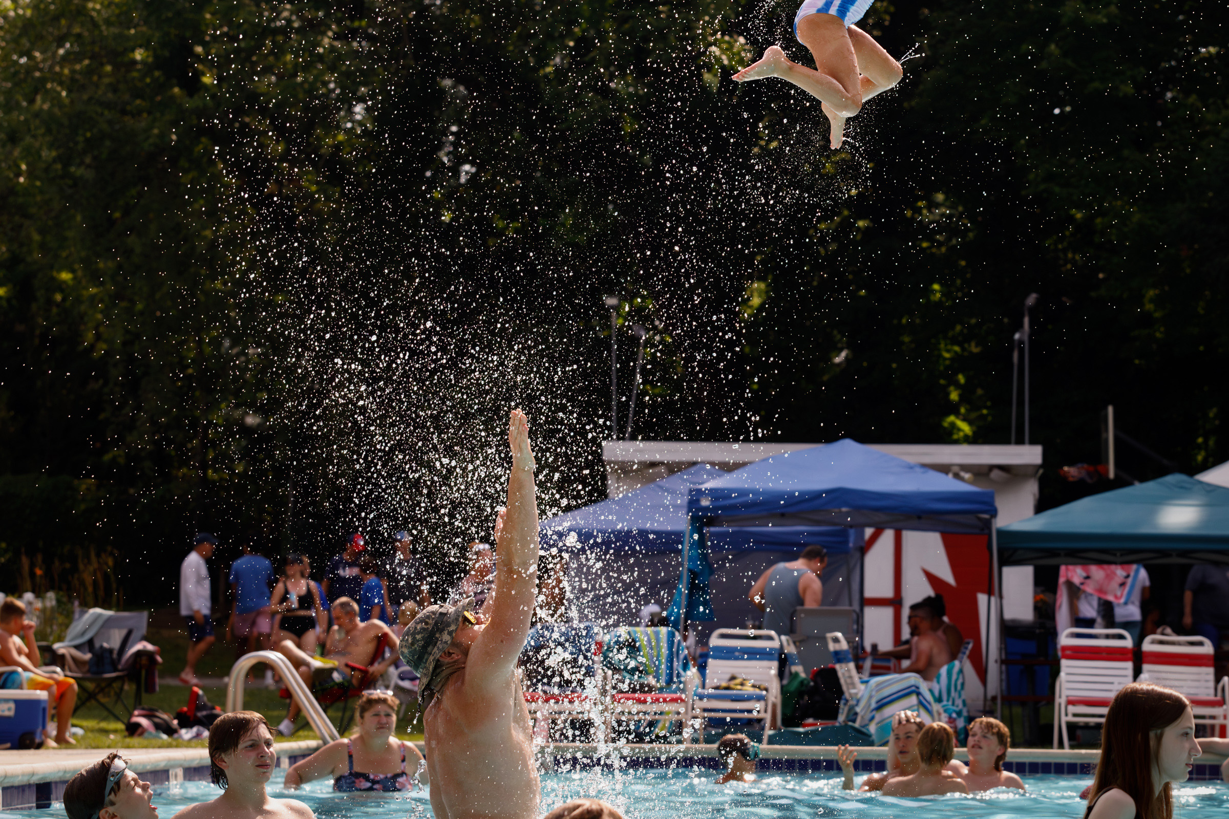 Father tosses his child high into the air above a crowded outdoor swimming pool as water sprays upward.