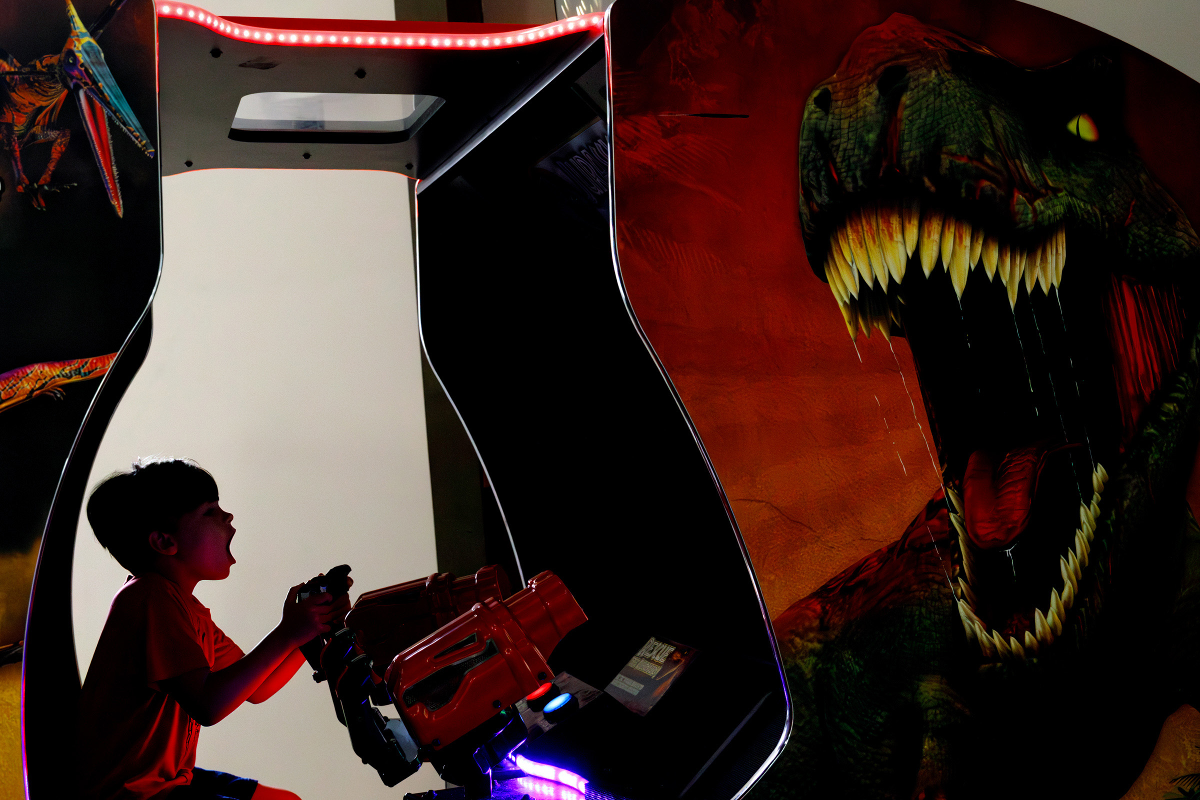 Child, mouth agape, plays a dinosaur-themed arcade game beside the image of a roaring Tyrannosaurus rex.